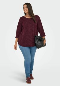 Top maroon con texture e maniche a tre quarti, abbinato a jeans skinny blu e sneakers borgogna. Una borsa nera aggiunge un dettaglio di contrasto.
