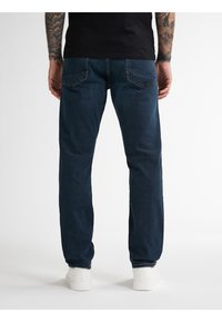 Vaqueros de mezclilla azul oscuro con un corte recto, que presentan bolsillos traseros con detalles de costura. Combinados con zapatillas blancas.