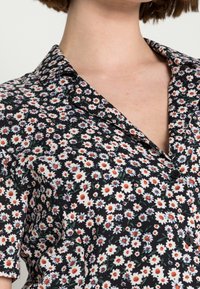 Blusa floreale con sfondo nero, caratterizzata da piccoli fiori bianchi, arancioni e blu, colletto e chiusura frontale con bottoni.