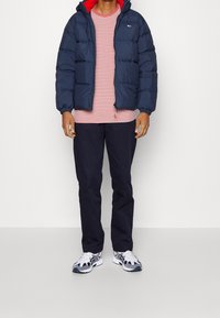 Hombre vestido con una chaqueta acolchada azul marino sobre una camiseta a rayas rojas y blancas, pantalones azul marino y zapatillas blancas con detalles azules, de pie sobre un suelo blanco.