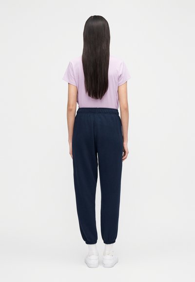 Kvinne med langt mørkt hår iført lys lilla t-skjorte, marineblå joggebukser, hvite sokker og hvite joggesko, stående mot en ensfarget hvit bakgrunn.