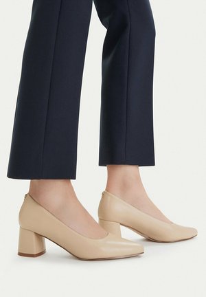 Par de zapatos beige con tacón bajo y cuadrado, usados con pantalones negros recortados, mostrando el perfil lateral y los tobillos sobre un fondo liso.