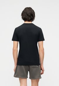 Άτομο με σκούρα, κυματιστά μαλλιά που φοράει απλό μαύρο κοντομάνικο μπλουζάκι και γκρι σορτς, στέκεται με την πλάτη σε ανοιχτό γκρι φόντο.