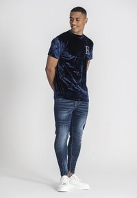 Námořnické sametové tričko s krátkým rukávem a logem K, spárované se slim-fit modrými džíny s opotřebeným vzhledem a bílými teniskami.