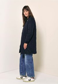 Cappotto imbottito blu navy con silhouette dritta, maniche lunghe, tasche laterali e chiusura con zip, abbinato a jeans a gamba larga blu chiaro e sneakers.