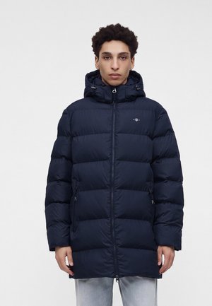 Junge Person mit kurzen lockigen Haaren trägt eine dunkelblaue wattierte Winterjacke mit Kapuze und Frontreißverschluss und steht vor einem einfarbigen hellen Hintergrund.