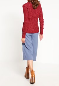 Pull texturé rouge, pantalon large bleu clair, et bottines à talons marron. Le pull est cintré ; le pantalon a une coupe ample.