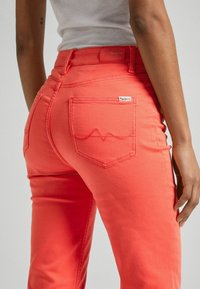 Jeans de mezclilla coral con un diseño ajustado, que presentan un bolsillo trasero con un patrón de olas y una etiqueta de logo. Textura suave con un ligero estiramiento.