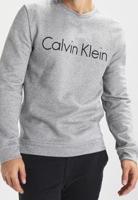 Sudadera gris con un acabado texturizado, que presenta un logotipo negro de "Calvin Klein" en el pecho, cuello redondo y puños acanalados.