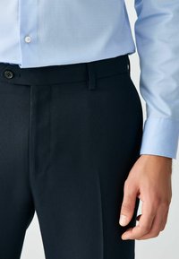 Marineblauwe broek met een gestructureerde afwerking, voorzien van een platte voorkant, riemlussen en een enkele knoopsluiting, gecombineerd met een lichtblauw shirt.