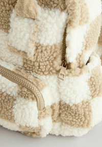 Sac à dos en polaire texturée beige et crème avec un motif à carreaux ; il dispose d'une fermeture éclair en plastique avec une tirette rectangulaire.