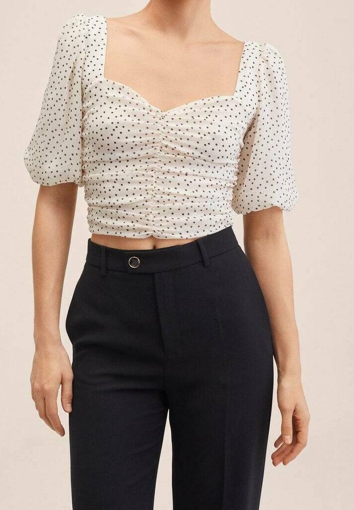 Femme portant un crop top blanc à manches courtes bouffantes à pois et un pantalon noir taille haute, debout devant un fond beige.