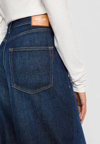 Nærbilde av person som har på seg mørkeblå Replay-jeans med synlig baklomme og lærmerke over midjebåndet.