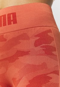 Legginsy sportowe o wysokim stanie, wykonane z teksturowanej tkaniny w wzory. Kolor: koralowy czerwony z subtelnym wzorem w kamuflaż i prążkowanym pasem.