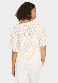 Top in pizzo color avorio con motivi floreali a intaglio, maniche a scallop e vestibilità comoda. La vista posteriore mostra un design a costine aperte.