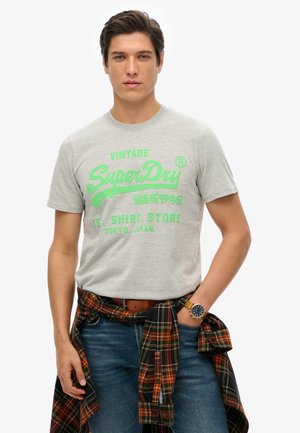 Szary T-shirt z krótkim rękawem z dużym zielonym grafikiem i napisem "SuperDry". Nosi go w połączeniu z niebieskimi dżinsami i czarną koszulą w kratę zawiązaną w pasie.