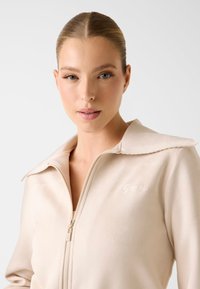 Veste beige zippée avec col montant, texture douce et logo brodé "Guess" sur le côté gauche de la poitrine.