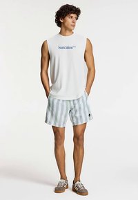 Camiseta blanca sin mangas con texto "Suncation", combinada con pantalones cortos a rayas en verde claro y blanco. Se lleva con zapatillas marrones.