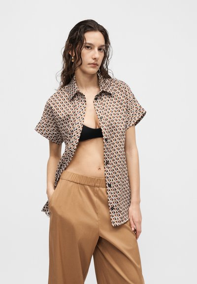 Γυναίκα με βρεγμένα μαλλιά φοράει κοντομάνικο πουκάμισο με γεωμετρικό σχέδιο ανοιχτό πάνω από μαύρο σουτιέν, συνδυασμένο με φαρδιά καφέ παντελόνια, πόζα με το χέρι στην τσέπη.