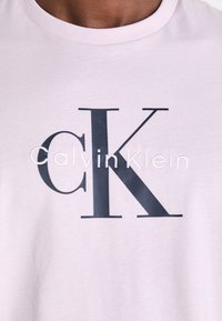 Svijetloružičasta pamučna majica s tamnosivim "CK" logotipom i izvezenim "Calvin Klein" ispod u kontrastnoj bijeloj boji.