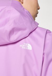 Lila, könnyű héjkabát kapucnival, amelyen a "The North Face" fehér logó található a hátsó részen. Sima, texturált anyag minimális részletezéssel.