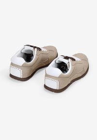 Zapatillas de ante beige con acentos blancos, suelas de goma marrón y cordones marrones. Presentan un diseño lateral texturizado y un detalle de costura en la parte trasera.