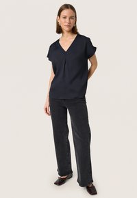 Top corto de manga corta azul oscuro con escote en V y detalle de pliegue, combinado con pantalones vaqueros de pierna ancha negros y zapatos marrones, sobre un fondo neutro.