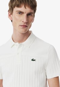 Vit pikétröja med ribbad textur, klassisk krage, knappslå och en grön logotyp på vänster bröst. Mjuk bomullstyg.