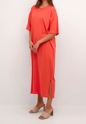 Femme vêtue d'une robe corail ample, manches courtes, longueur mollet avec une fente latérale et des sandales beiges à talon bloc ouvert, debout devant un fond uni.