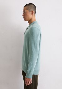 Jasnoniebieski sweter z dzianiny z kołnierzem, trzema guzikami, ściągaczami na rękawach i dole. Noszony z ciemnymi spodniami, widoczny z boku na neutralnym tle.