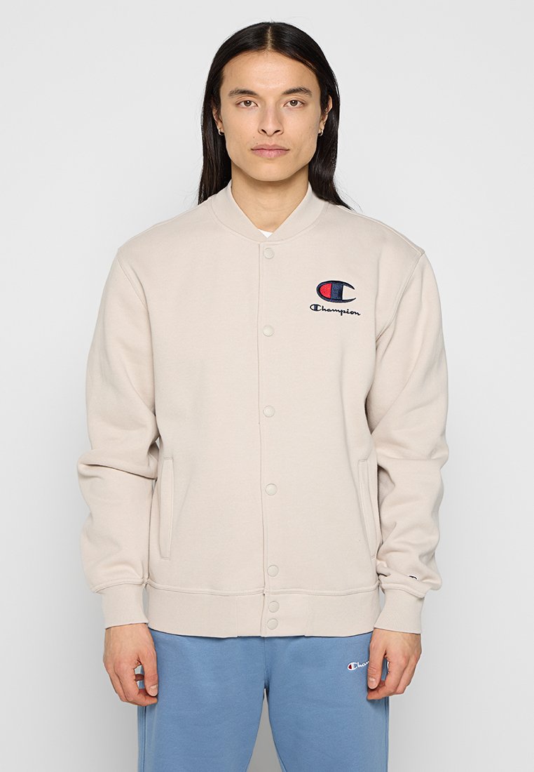 Giacca in pile beige chiaro con colletto rotondo, bottoni a pressione frontali, due tasche laterali e un logo blu e rosso sul lato sinistro del petto.