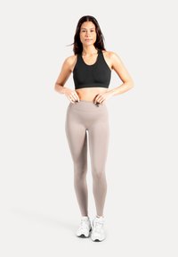Zwarte sportbeha met een ronde halslijn, gecombineerd met lichtbeige hoge taille leggings. Model draagt witte sportschoenen.