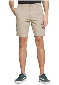 Shorts en coton beige à coupe droite, avec poches avant et arrière, ourlet retroussé, portés avec des baskets en daim vert.