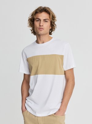 Giovane con capelli ricci biondi che indossa una maglietta bianca con una larga striscia orizzontale beige e pantaloni beige, con le mani nelle tasche.