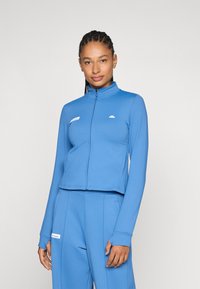 Kort blå zip-up jakke i strækstof med lange ærmer, ståkrave og logo detaljer på brystet og ærmet.