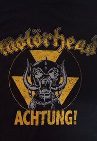 Camiseta negra con un gráfico amarillo que presenta un cráneo feroz con colmillos, detalles de cadenas y el texto "motörhead" y "ACHTUNG!" en letras negritas.