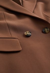 Brun kavaj med ett mjukt tyg, knappslå och två stora, runda knappar i sköldpaddsmönster med synlig sömnad.