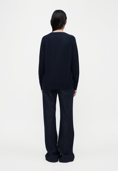 Άτομο με μακριά σκούρα μαλλιά που φοράει μπλε πουλόβερ ναυτικού τύπου και σκουρόχρωμα τζιν με φαρδιά μπατζάκια, στέκεται με την πλάτη προς το φωτεινό, απλό φόντο.