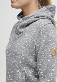 Sudadera gris hecha de tela suave, con cuello tipo cowl, botones decorativos y un patrón disperso de pequeños círculos azules y rosas.