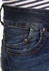 Tmavě modré džíny vyrobené z denimu s přiléhavým pasem a detailním šitím. Mají dvě zadní kapsy se měděnými knoflíky a texturovanou tkaninou.
