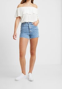 Off-shoulder vit topp med volanger, tillsammans med blå denimshorts med ett repetitivt Levi's logodesign, och vita sneakers.