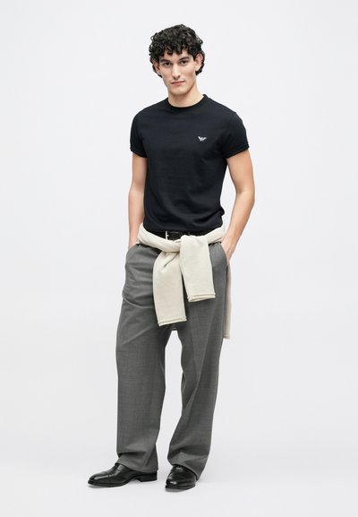 Juoda aptempianti marškinėliai su apskritu iškirpimu ir mažyčiu logotipu, derinami su šviesiai pilkomis plačiomis kelnėmis ir smėlio spalvos megztiniu, surištu ties liemeniu. Juodi batai.