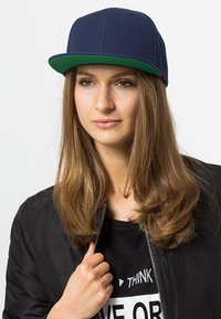 Marineblå baseballcap med grønn skygge, strukturert design, laget av stoff, brukt med en svart bomberjakke og grafisk t-skjorte.