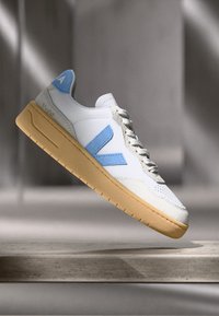 Basket blanche avec accents bleu clair et semelle beige. Présente une texture perforée, une languette texturée, et un logo sur le talon et sur le côté.