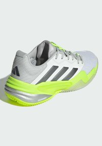 Zapatilla deportiva gris con parte superior de malla, brillantes acentos en verde neón, media suela plateada y tres franjas negras en el lateral. Suela texturizada.