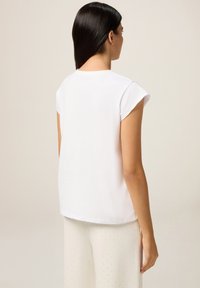 Mujer con cabello largo y oscuro, vestida con una camiseta blanca de mangas cortas y pantalones blancos con pequeños patrones de puntos, de espaldas frente a un fondo neutro.