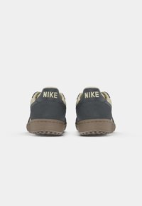 Le sneakers Nike presentano una parte superiore in suede grigio con un tallone color crema, due evidenti loghi "Nike" e una suola in gomma marrone texturizzata.