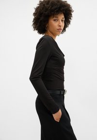 Črn zgornji del s long sleeves in prilegajočim se krojem, v-izrezom in gladko strukturo, v kombinaciji s črnimi hlačami, ki jih drži pas z kovinsko sponko.