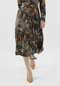 Květinové košilové šaty s tmavězeleným podkladem a přírodními odstíny, dlouhými rukávy a splývavým siluetam. Béžové vysoké podpatky.