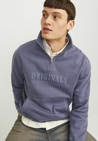 Pullover viola in pile con colletto a mezza zip, tasca frontale e logo in rilievo "ORIGINALS JACK & JONES". Indossato sopra una camicia bianca.